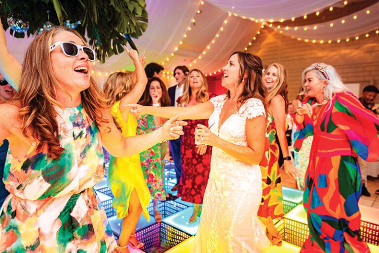Guests dance on a lit-up floor under string lights, including a bride in a lace gown and a woman in sunglasses.